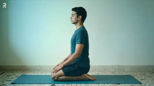 Young man kneeling in Vajrasana (Thunderbolt Pose) on a yoga mat with eyes closed and hands on his knees, a calming yoga pose for nightfall